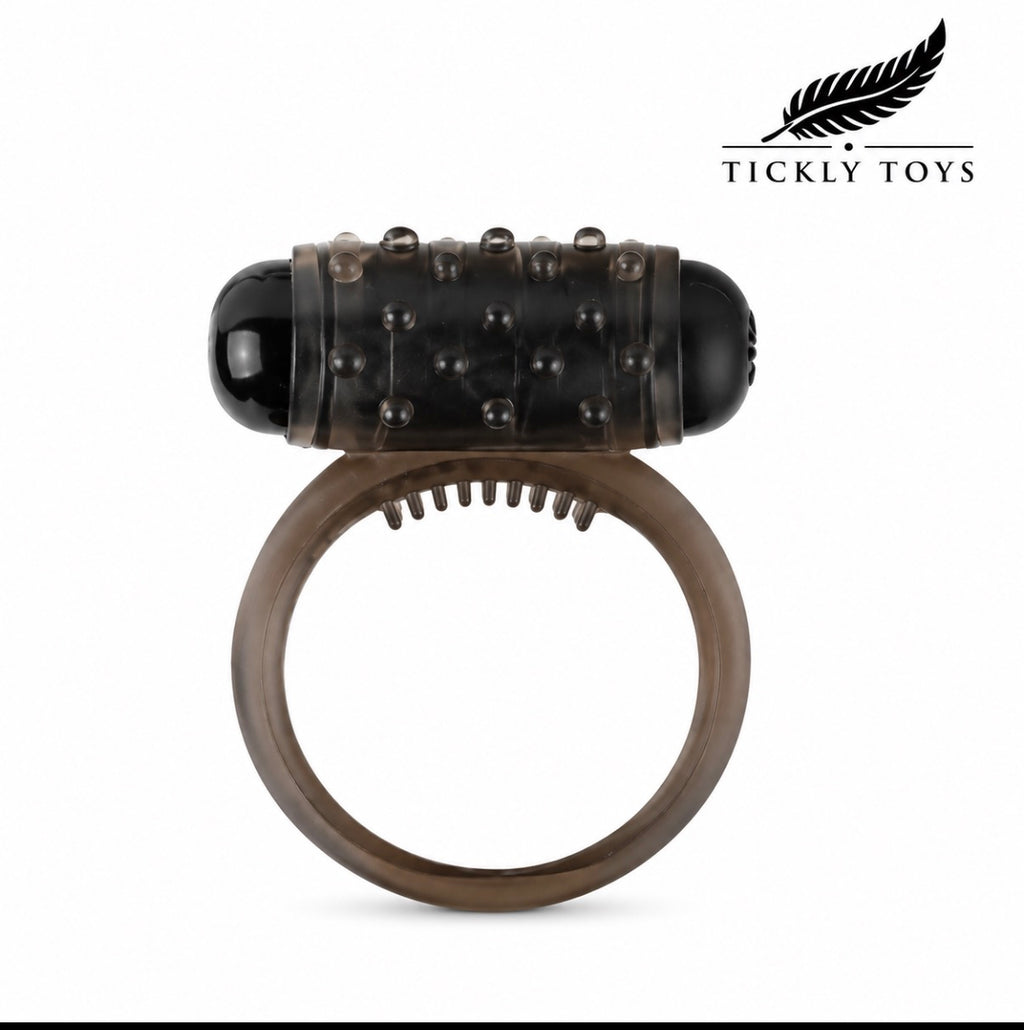 An adult novelty device featuring a translucent dark brown silicone ring with a textured black studded cylinder on top, shown against a plain white background with the Tickly Toys brand logo in the top right corner.