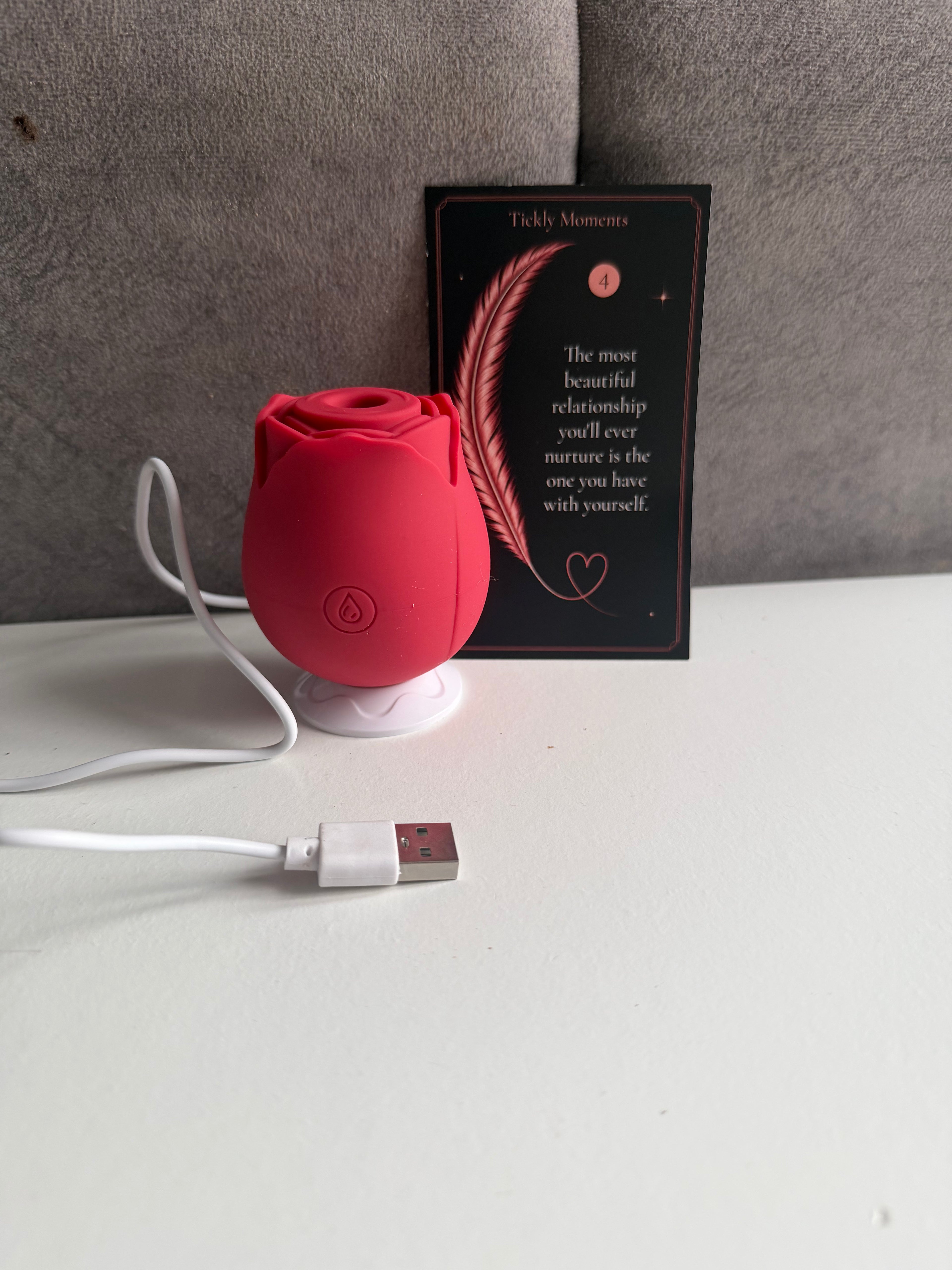 A small rose-shaped pink USB device on a white charging base, shown with a USB cable on a white tabletop in front of a gray upholstered chair. A black card behind it reads 'Tickle Moments' with a feather and quote.